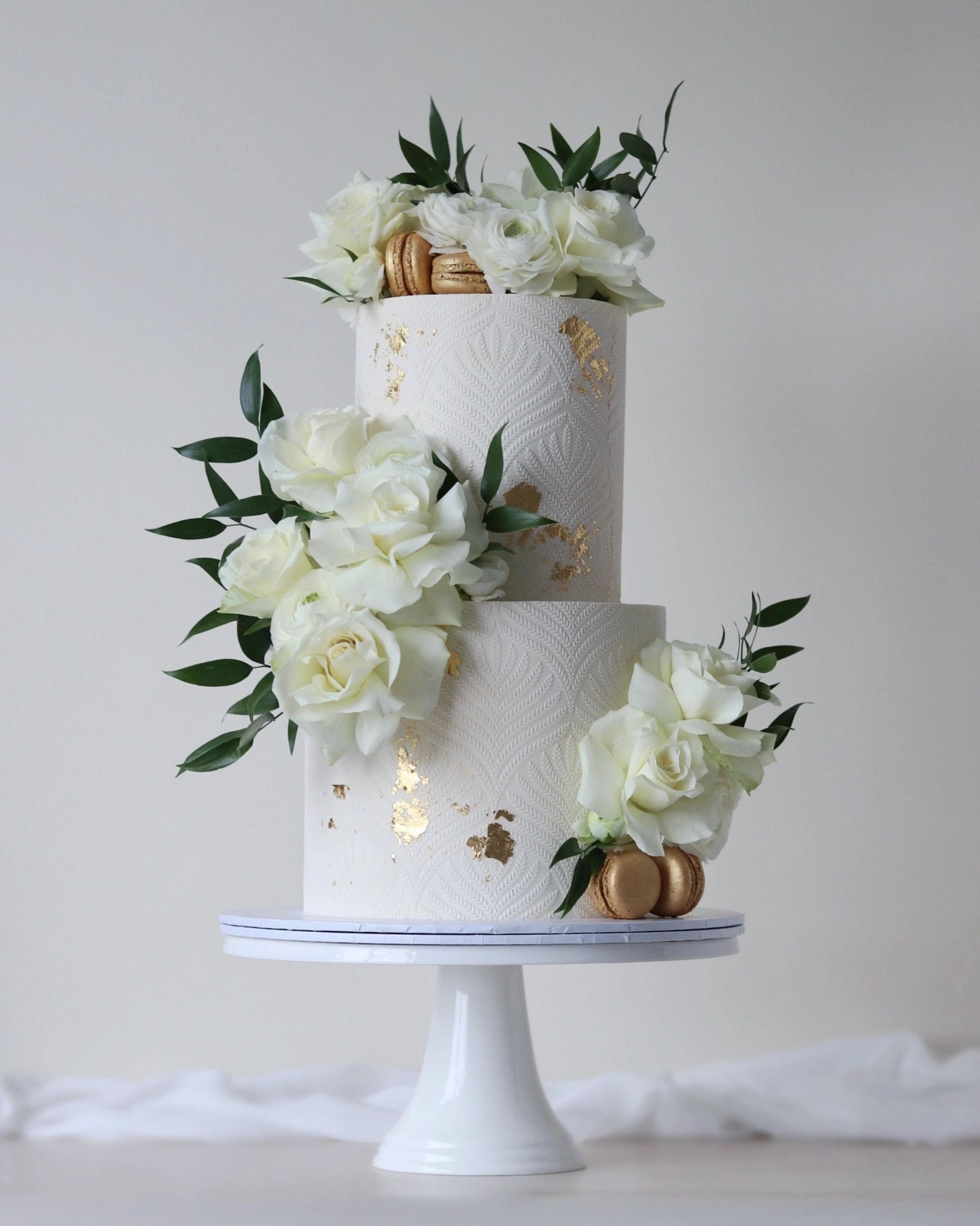 A modern wedding cake in ivory and gold. Featuring delicate all-around stencil overlay, fresh flowers, and gold accents. 