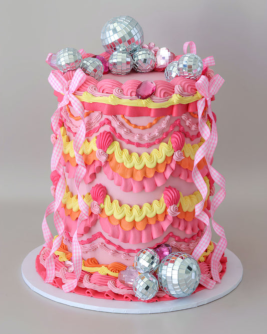 A tall vintage Lambeth birthday cake named Disco Diva, decorated with intricate pink, orange, and yellow ruffles. The cake features pink and white gingham checkered bows draped down the sides, large pink gems, and clusters of miniature silver mirror disco balls.